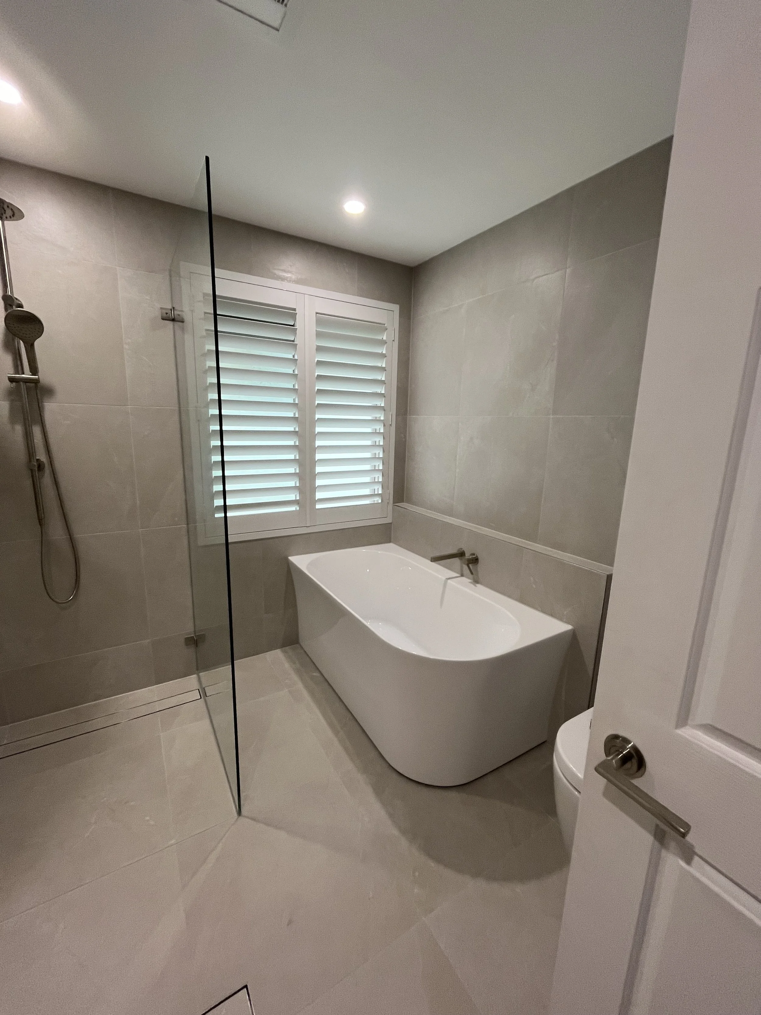 Modern bathroom with a white bathtub, gray tiled walls, a window with white shutters, and a glass shower partition.