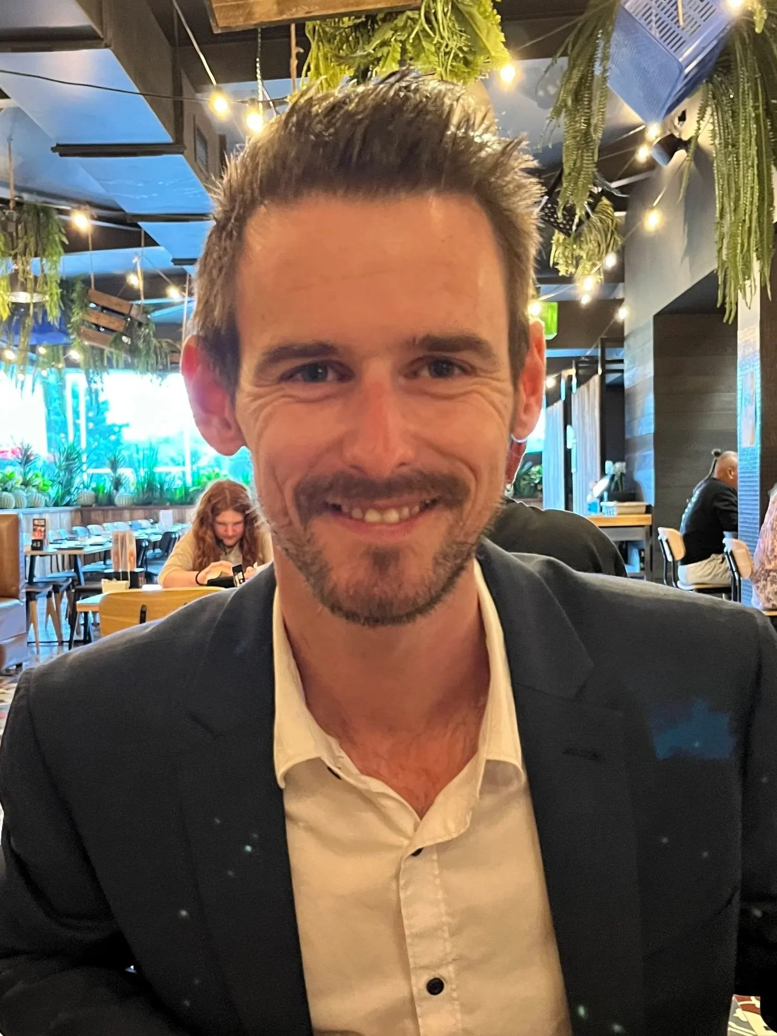 A smiling man with brown hair and a beard, dressed in a black suit and white shirt, sits in a restaurant with hanging plants, string lights, and other diners in the background.