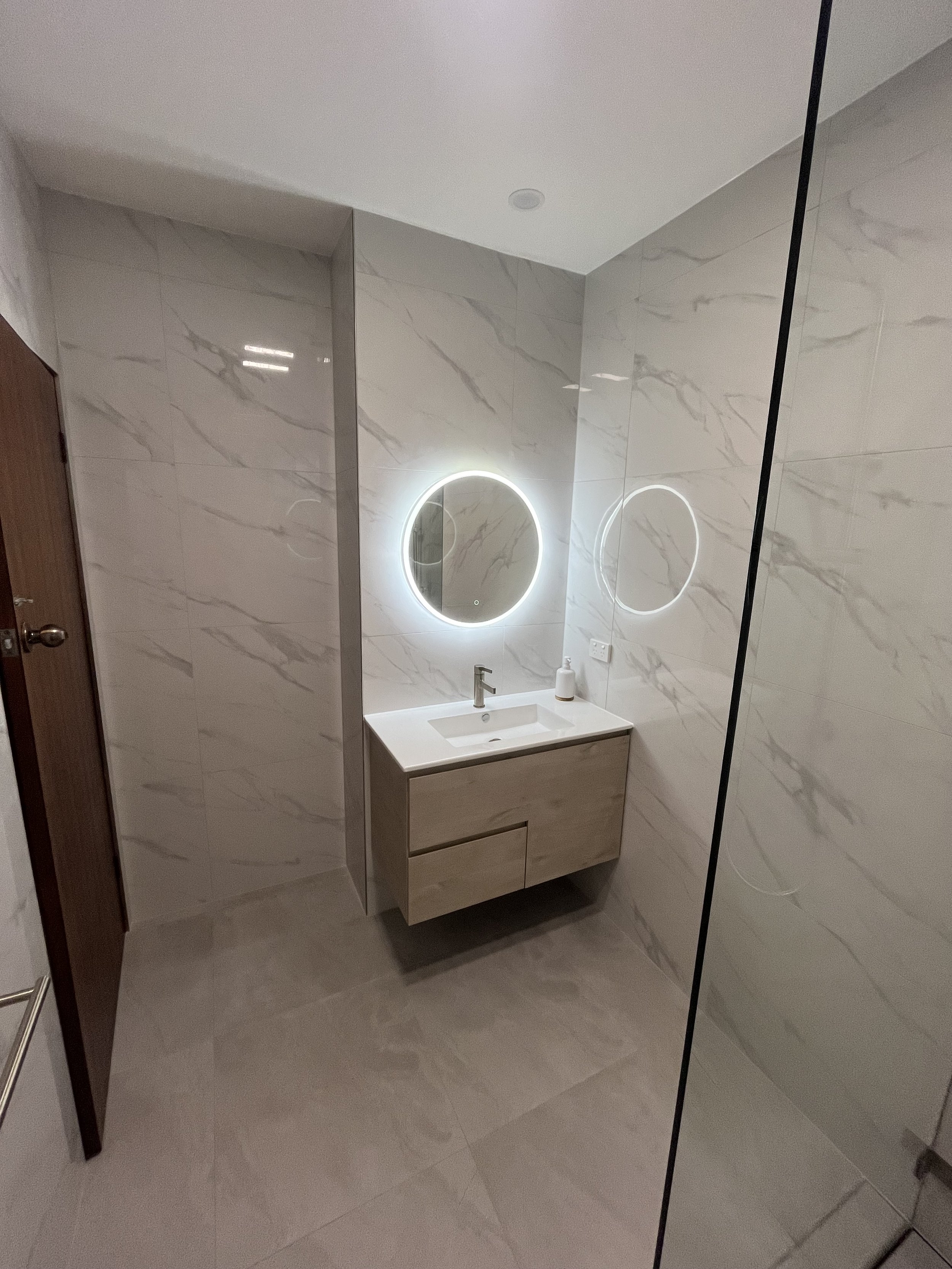 Modern bathroom with marble walls, a backlit circular mirror, a small white vanity with a sink, and a soap dispenser. There is a wooden door on the left and a dark divider or wall on the right.