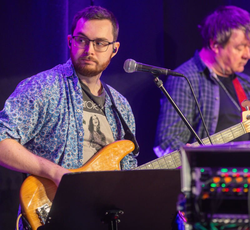 Two men playing guitars on stage with microphones and audio equipment, purple stage lighting.