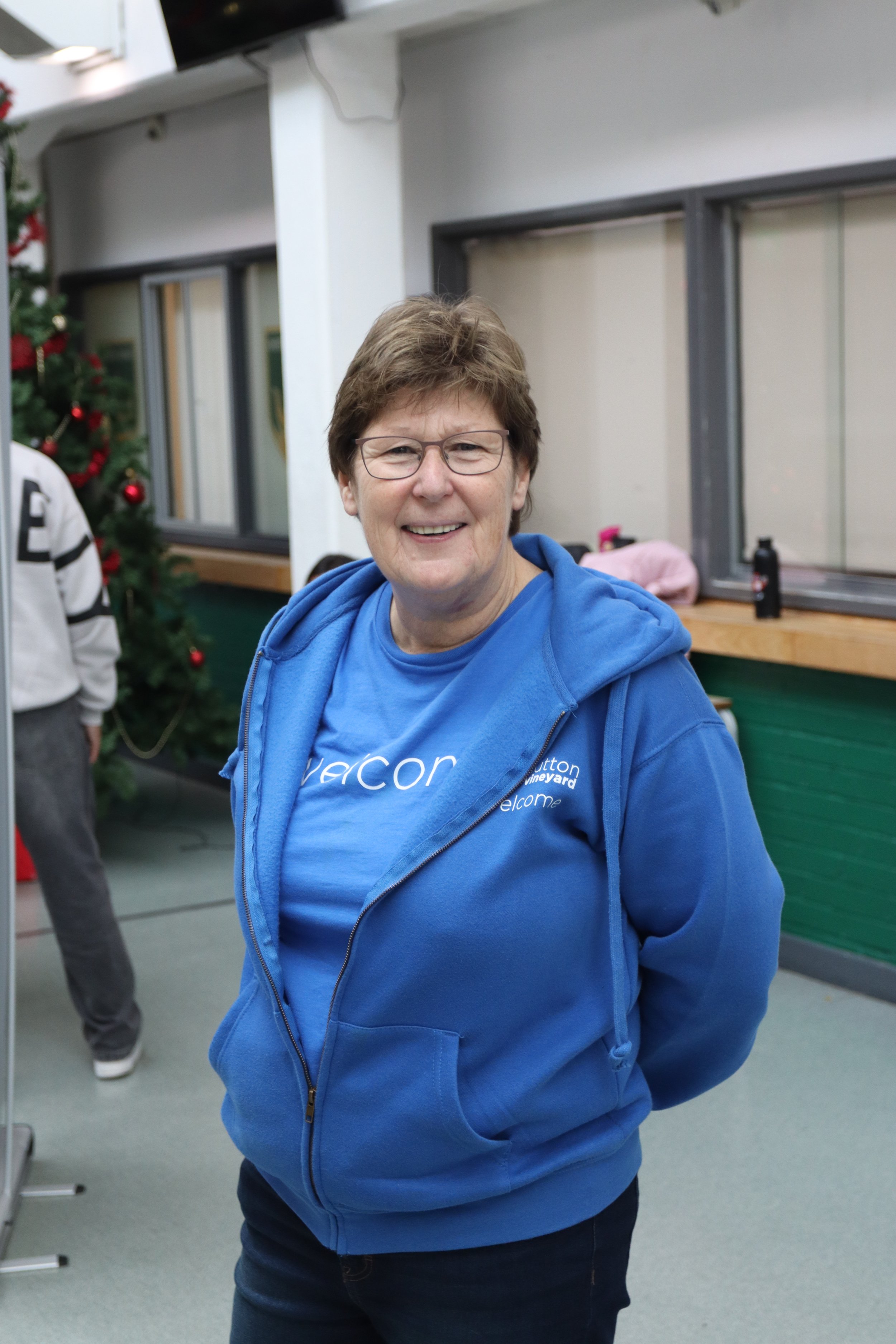 Smiling woman wearing glasses and a blue zip-up hoodie in an indoor setting with windows and a decorated Christmas tree in the background.