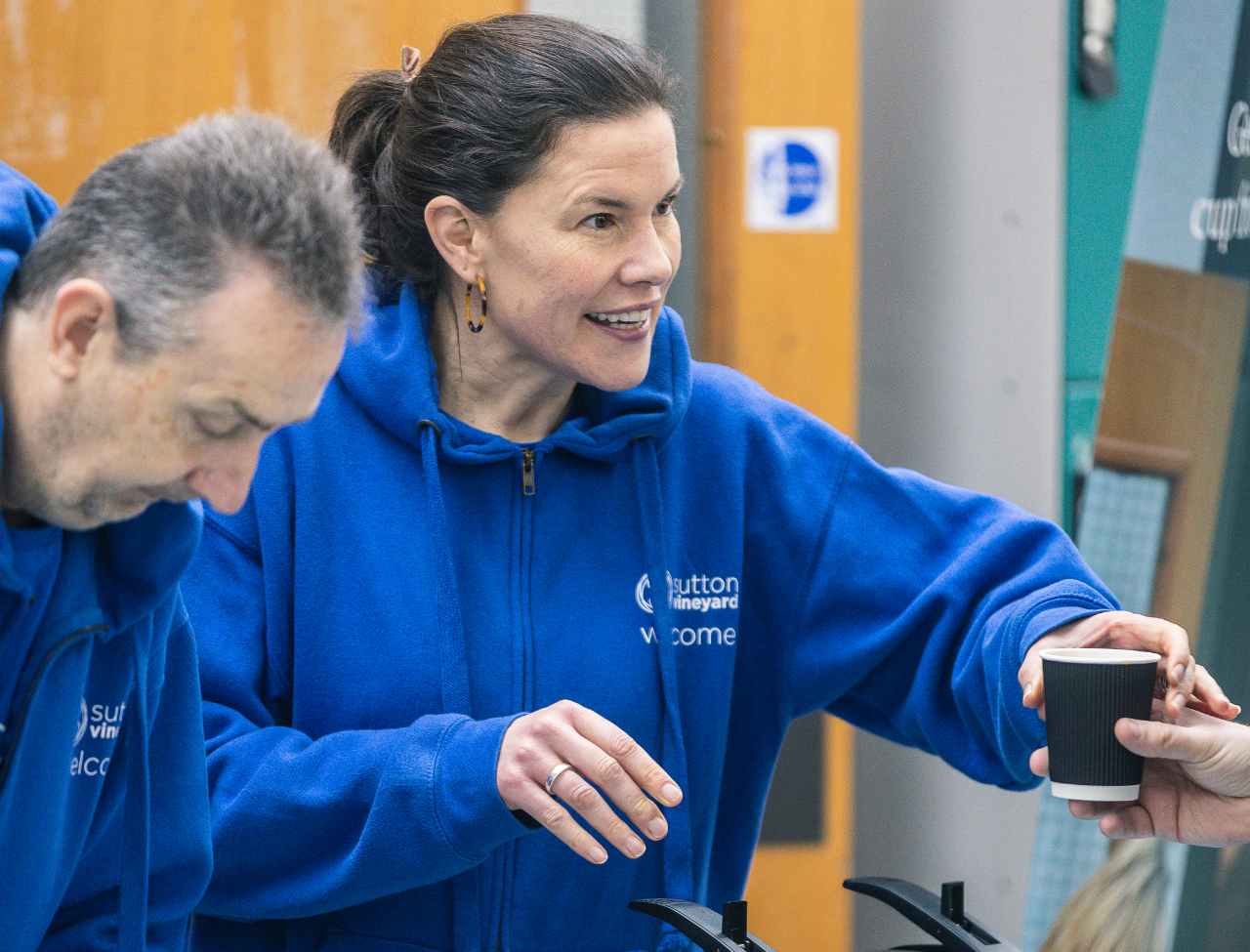 A woman in a blue hoodie is handing a black paper coffee cup to someone. She is smiling and standing next to a man, also wearing a blue hoodie, in an indoor setting with wooden walls.