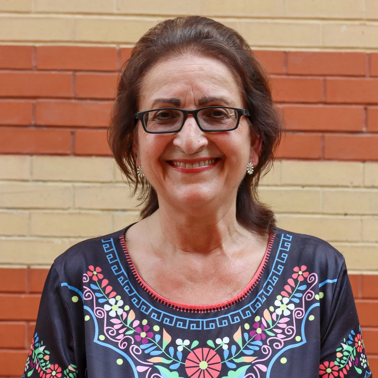 A smiling woman with short brown hair, wearing glasses, earrings, and a colorful embroidered black top, standing in front of a brick wall.