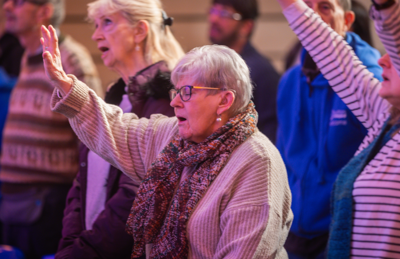 Older woman with glasses and a patterned scarf raising her hand, surrounded by other people at an event.