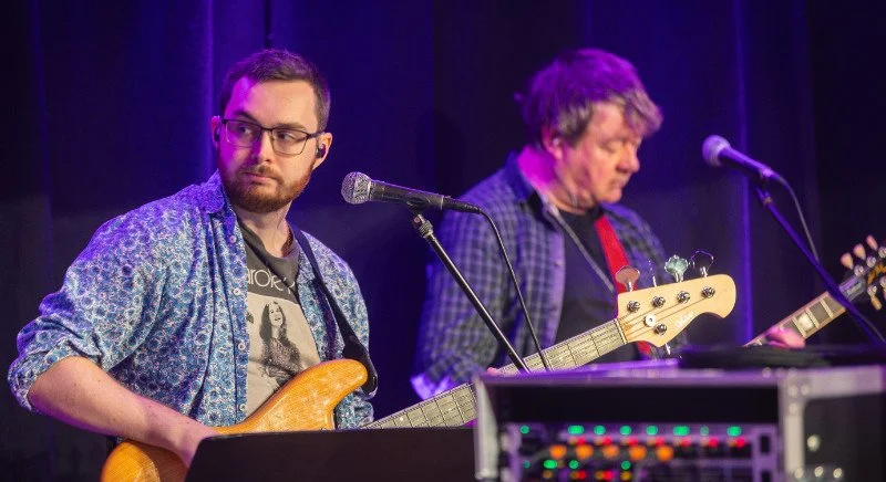 Two male musicians on stage with guitars, one with glasses in a patterned shirt, the other with longer hair in a plaid shirt, standing in front of microphones with a dark curtain background.