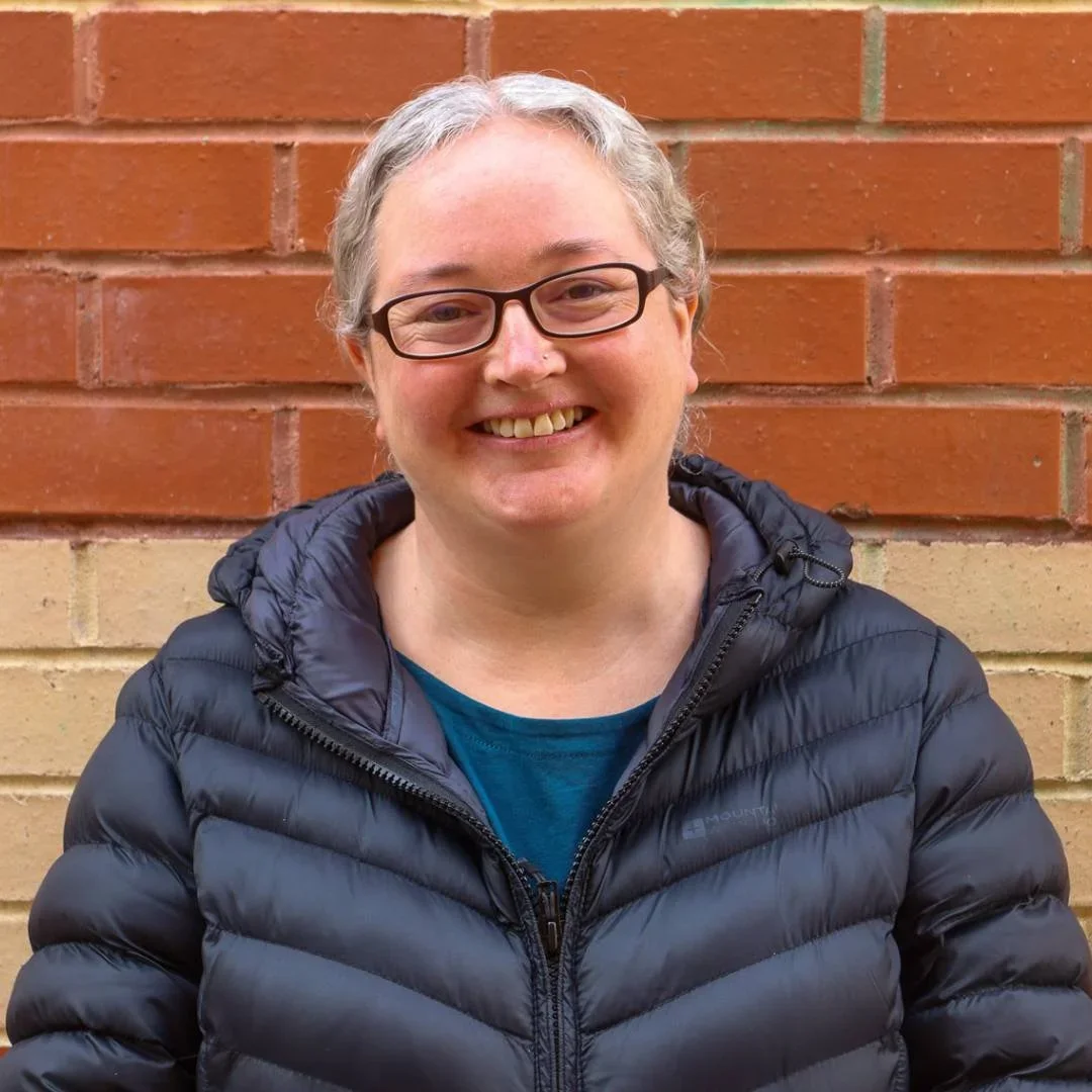 Smiling woman with glasses, gray hair, wearing a black puffer jacket, standing in front of a brick wall.