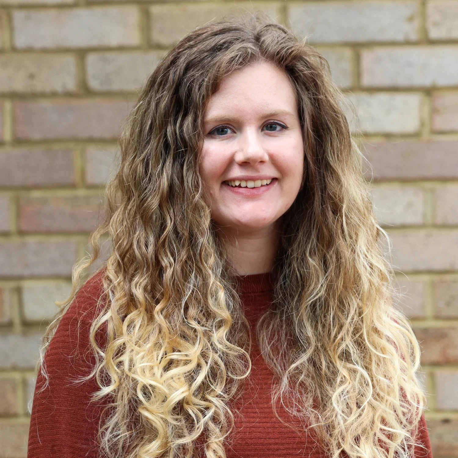 A young woman with long, curly blonde hair and blue eyes, smiling at the camera, wearing a rust-colored sweater, standing in front of a brick wall.