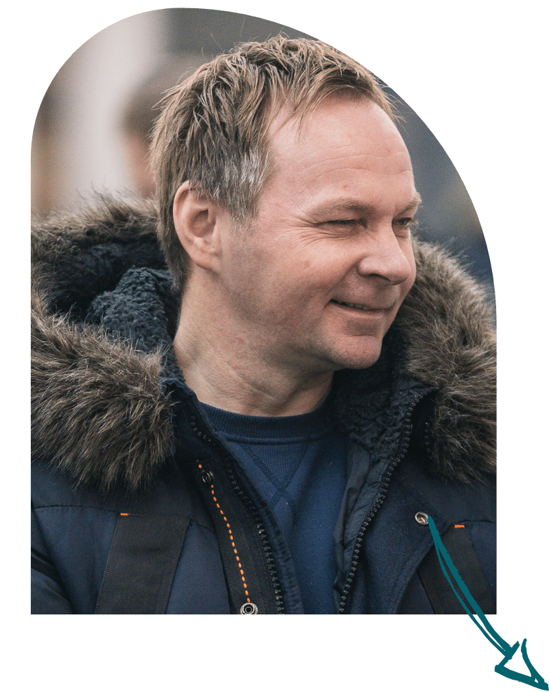 A smiling man with short, light brown hair wearing a dark winter coat with a fur-lined hood and a blue shirt underneath.