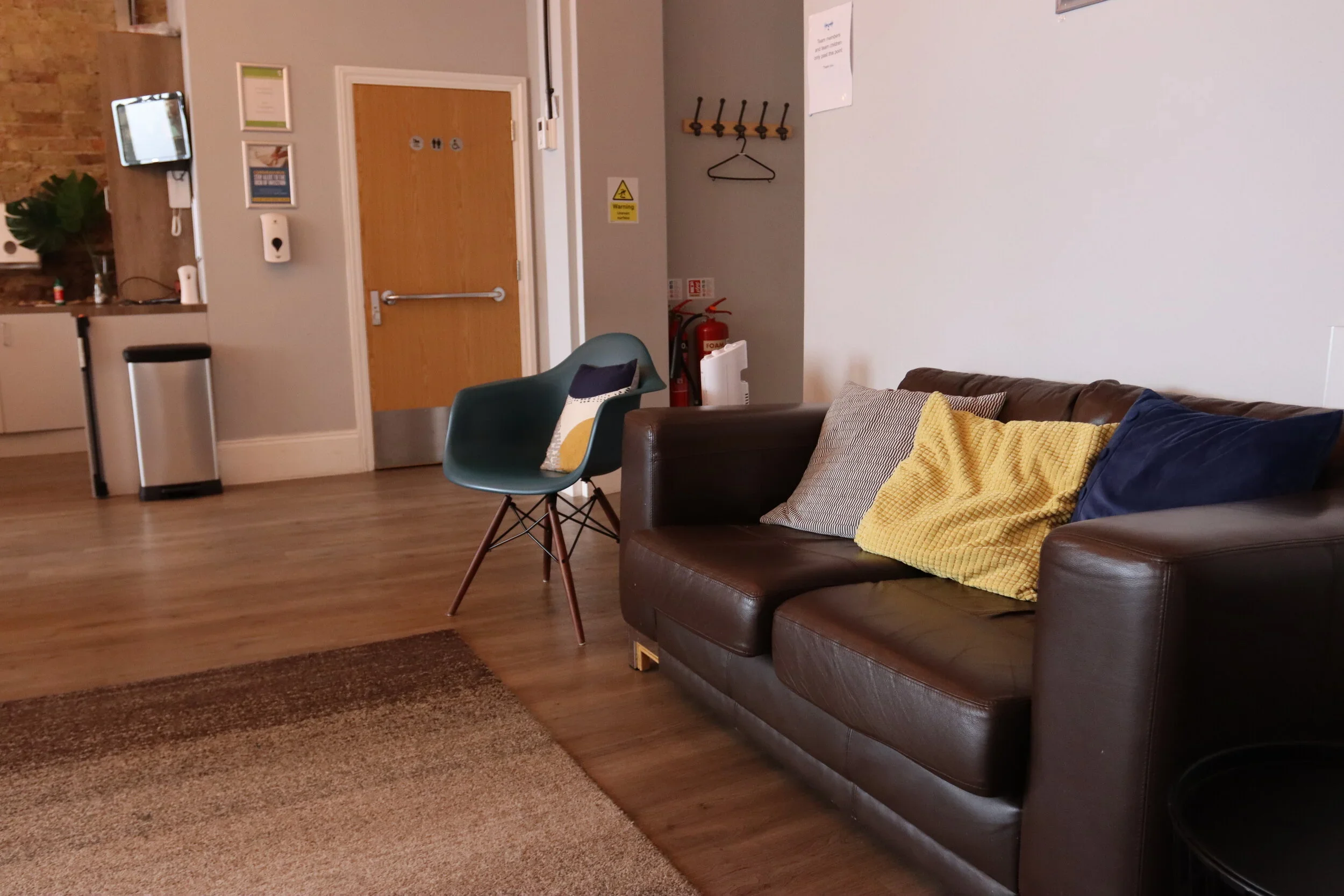 Living room with a brown leather sofa with pillows, a green chair with a pillow, a wooden floor, a beige rug, and a wall with a coat rack and fire extinguishers.