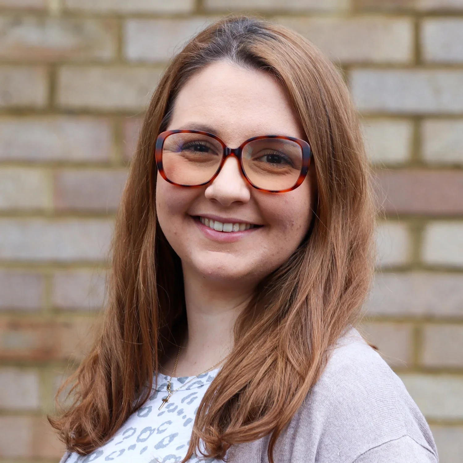 A young woman with long red hair, wearing large tortoiseshell glasses, smiling, in front of a blurred brick wall.
