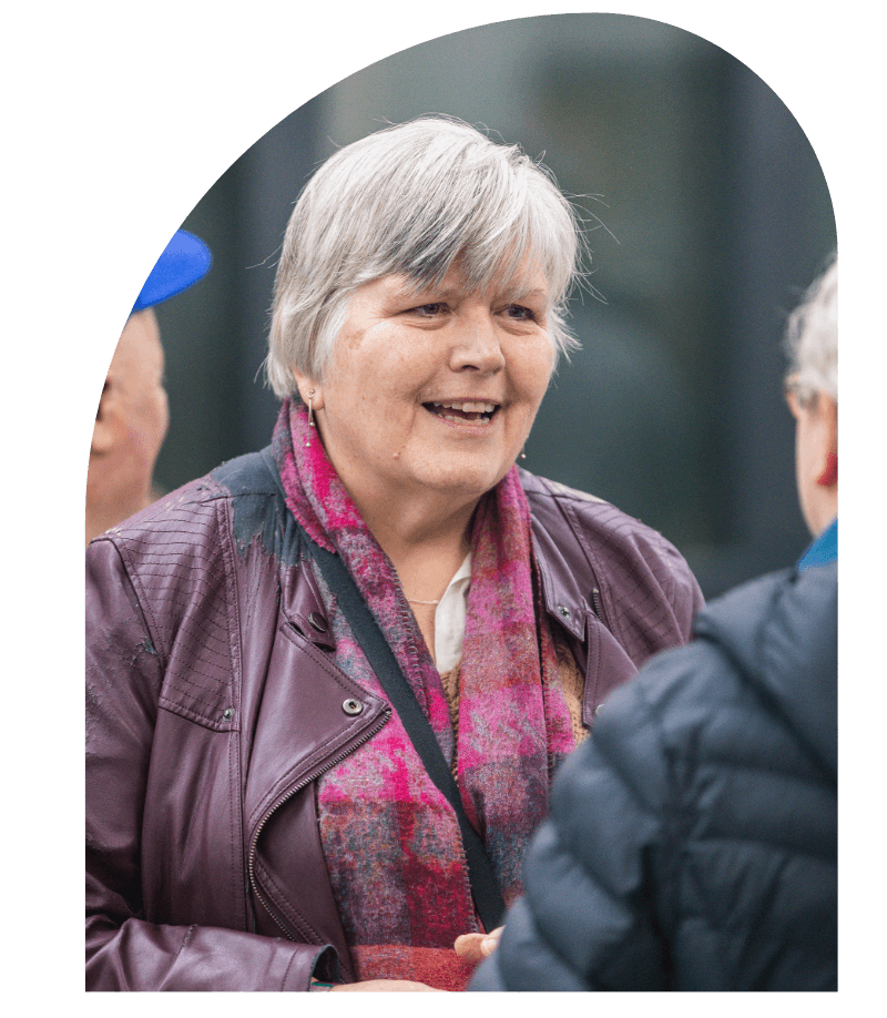 A smiling middle-aged woman with short gray hair talking to another person, wearing a purple jacket and pink scarf, outdoors in a social setting.