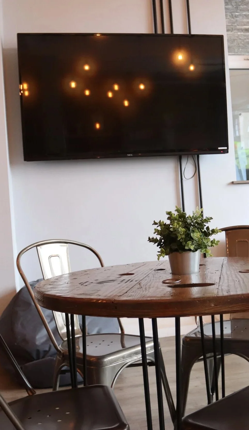 Interior of a cafe with a wooden table, metallic chairs, a potted plant, and a large television mounted on the wall.