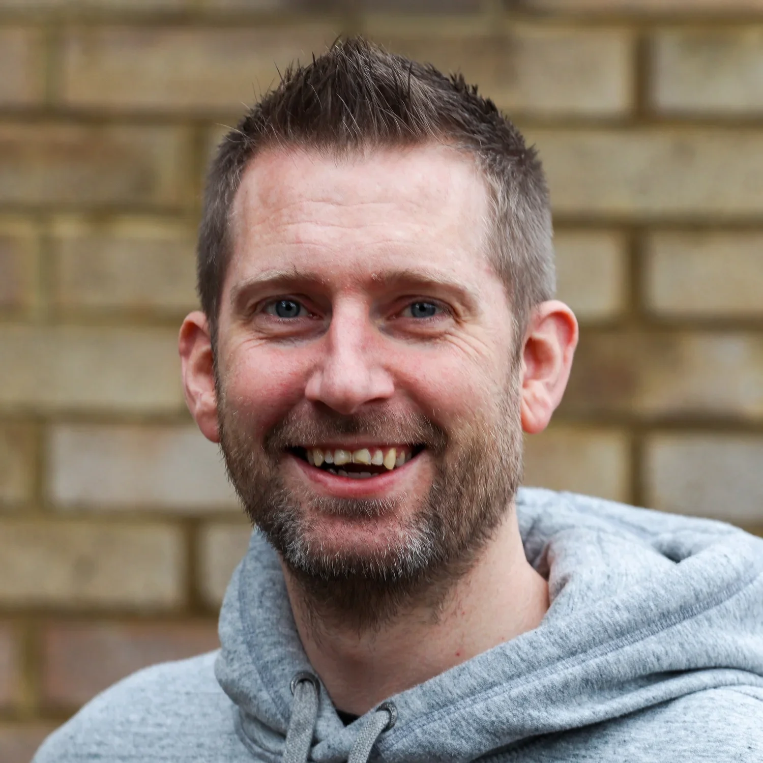 Close-up of a smiling man with short brown hair and a beard, wearing a gray hoodie, standing against a brick wall background.