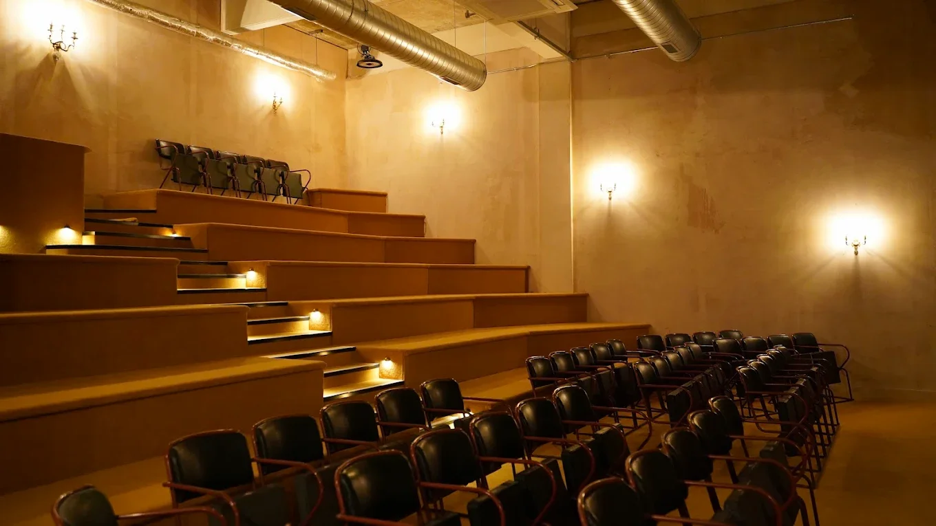 An empty auditorium-style theater with rows of black chairs and tiered seating, illuminated by wall sconces and small lights along the steps.