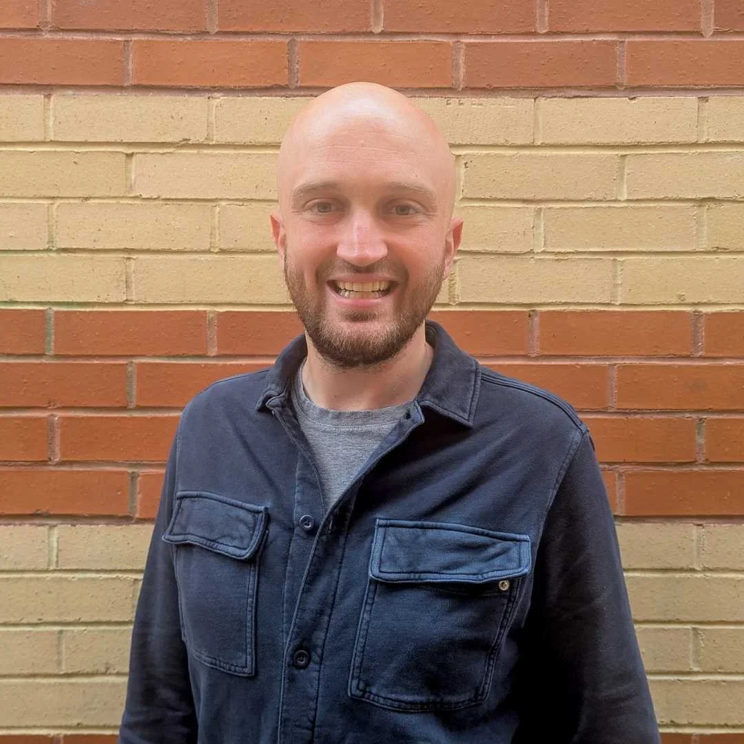 A smiling man with a bald head, beard, and light skin wearing a dark denim jacket and gray shirt, standing in front of a multicolored brick wall.