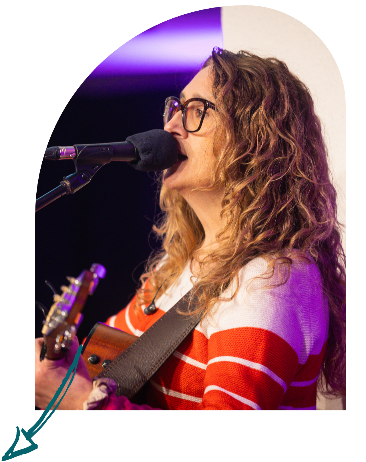 A woman with curly hair and glasses singing into a microphone while playing an acoustic guitar, wearing a red and white striped sweater.