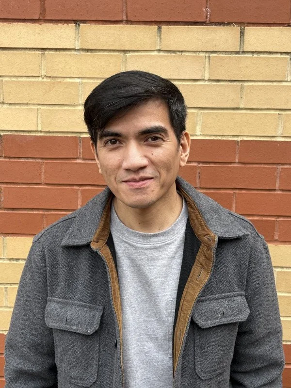 A man with black hair wearing a gray jacket over a light gray shirt, standing in front of a yellow and red brick wall.