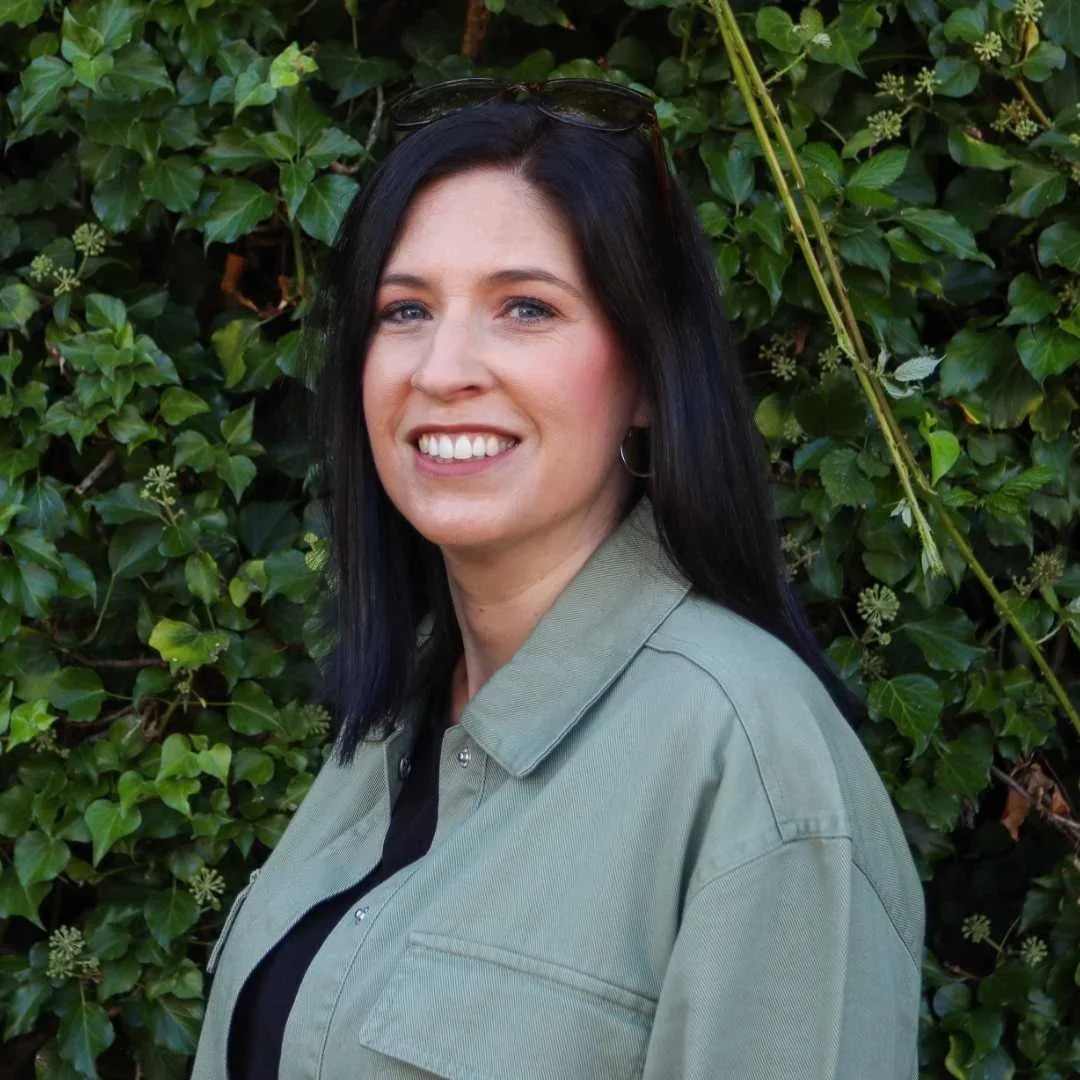 A woman with long black hair, wearing a green shirt and sunglasses on her head, smiling outdoors in front of a leafy green hedge.