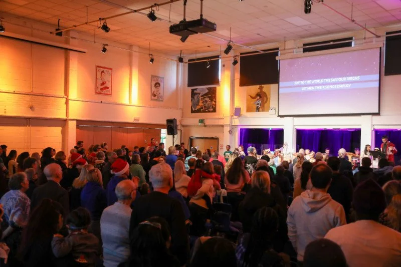 Crowd of people gathered in a large indoor space watching a stage performance with a large screen displaying music lyrics.