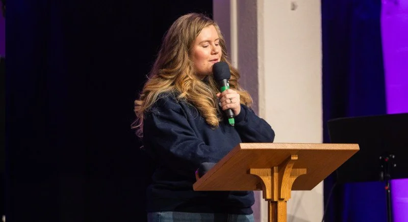 A woman with long wavy hair holding a microphone and speaking at a wooden podium on a stage with purple and black background.