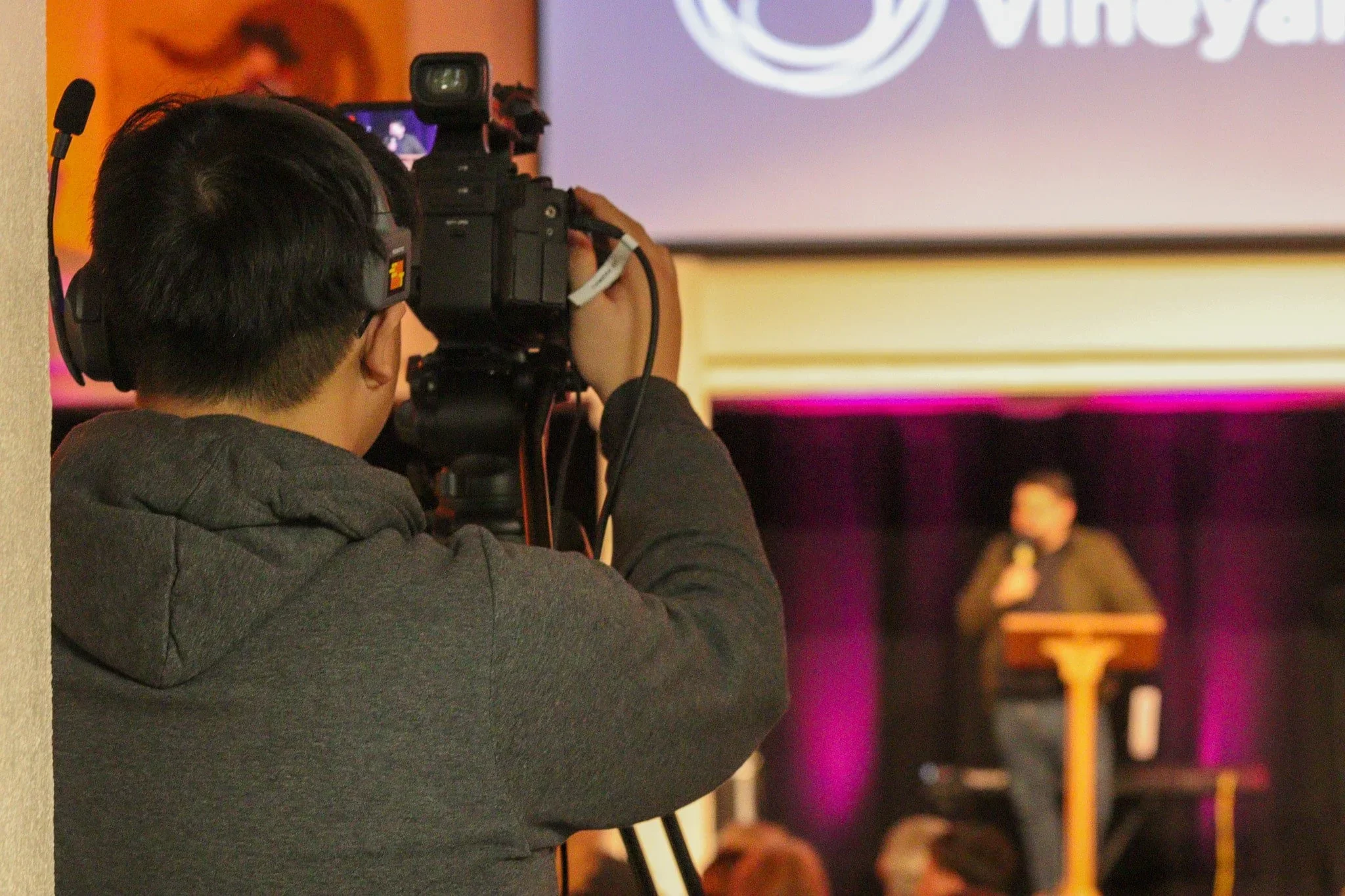 A person with short, dark hair is using a professional camera to film or photograph a person standing on a stage. The background shows a large screen with some text and a logo, with purple and orange lighting creating an indoor event or presentation setting.