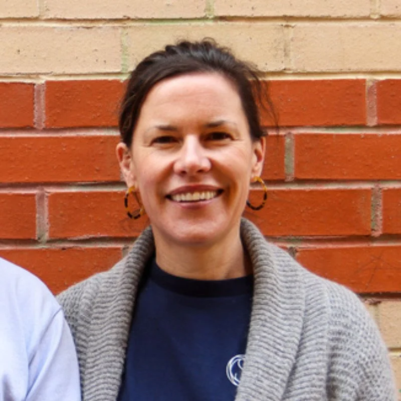 A smiling woman with dark hair, wearing hoop earrings, a dark blue shirt, and a gray cardigan, standing in front of a brick wall.