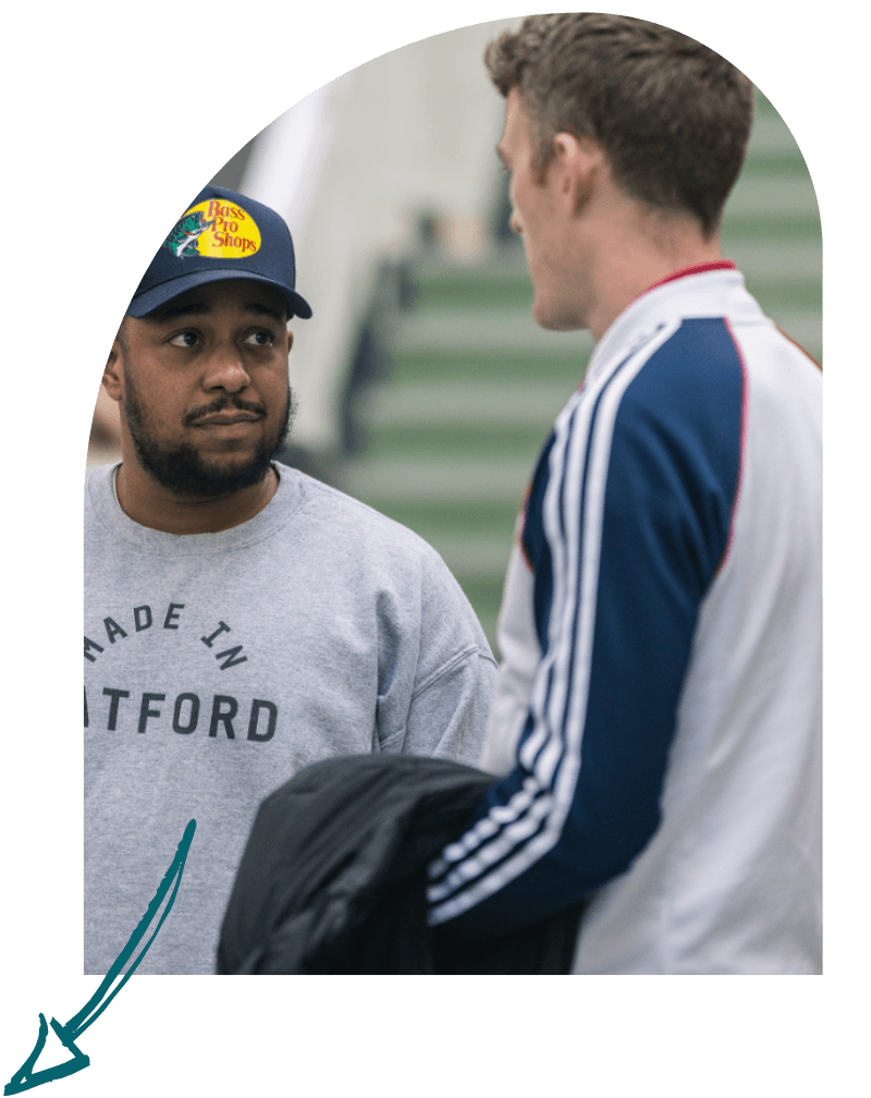 Two men talking outdoors, one wearing a jacket and the other a sweatshirt and cap, with green stairs in the background.