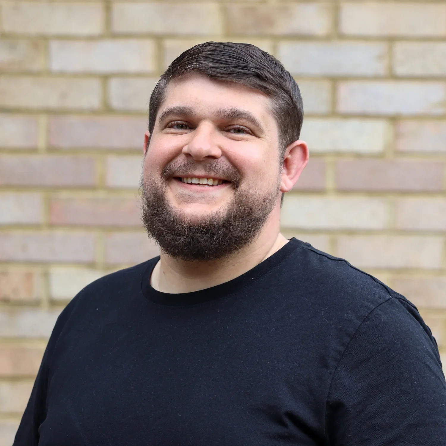 A smiling man with dark hair and a beard wearing a black T-shirt, standing in front of a brick wall.