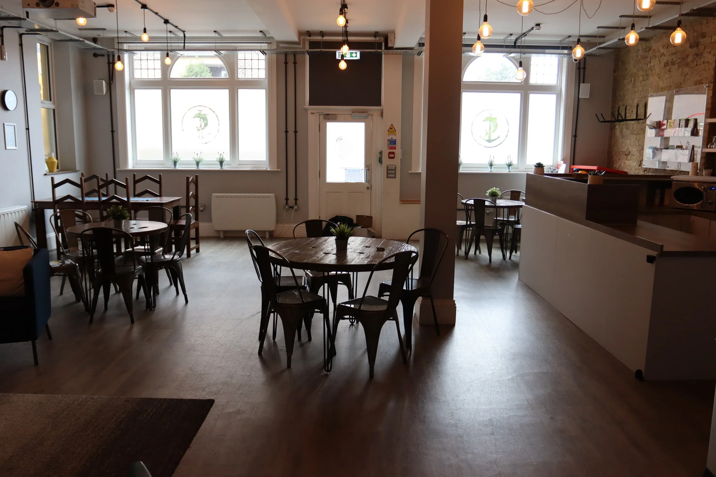 Interior of a cafe or restaurant with wooden tables and chairs, large windows with potted plants, exposed hanging light bulbs, and a brick wall with shelves and a microwave.
