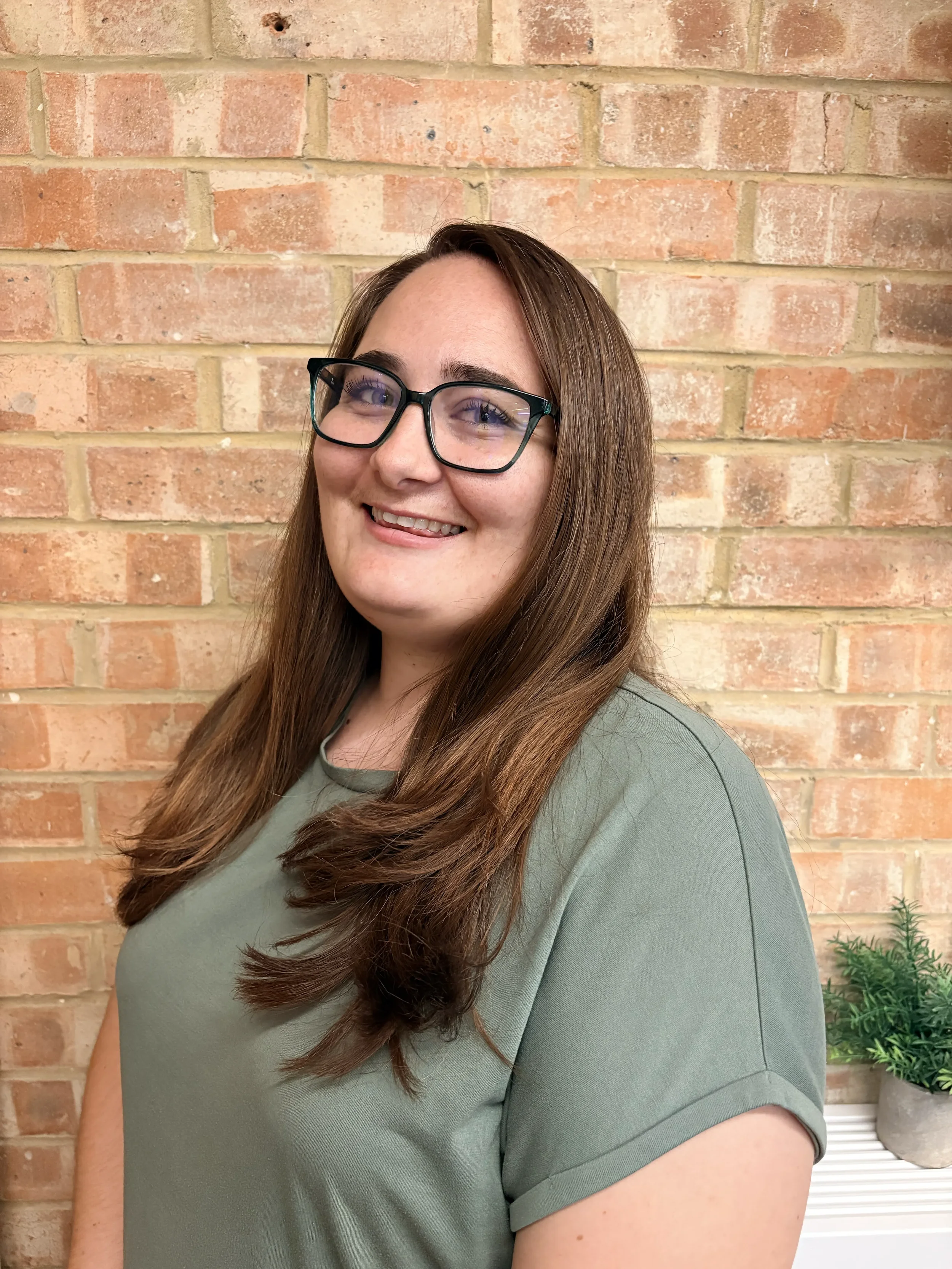 Close-up of a smiling woman with long brown hair and black glasses, standing against a brick wall with a small green plant in a pot beside her.