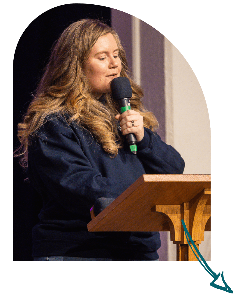 A woman with long, curly auburn hair speaking into a microphone at a wooden podium.