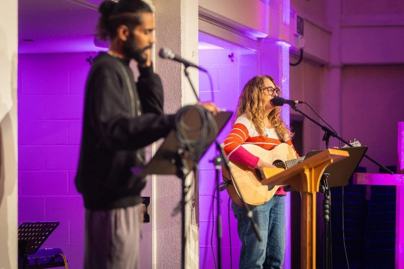 A woman with glasses playing an acoustic guitar and singing into a microphone on a stage with purple lighting, while a man with a ponytail reads from a script on a stand.