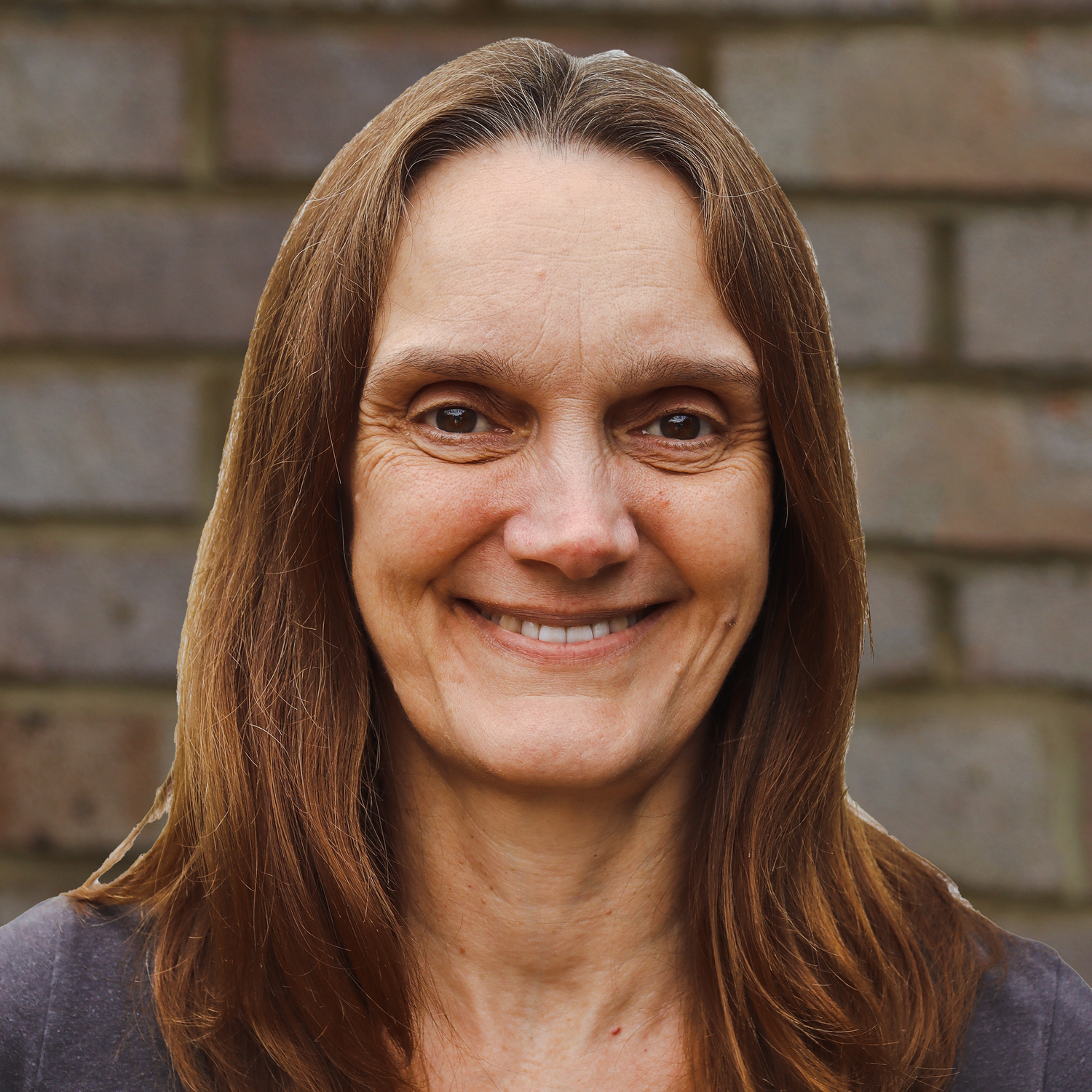 Close-up portrait of a smiling woman with long brown hair, in front of a brick wall.