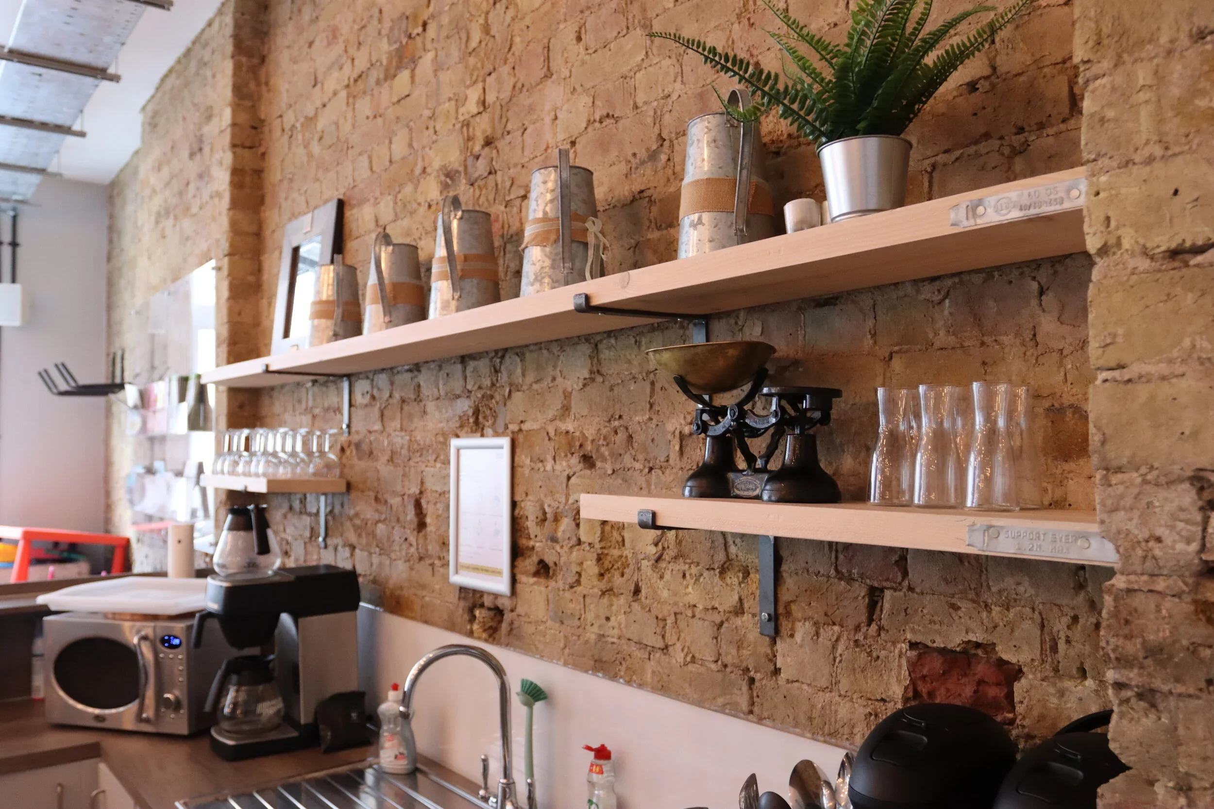 A kitchen with a rustic exposed brick wall. There are two wooden shelves holding metallic and ceramic kitchen items, including pots and pitchers. Below the shelves, there is a countertop with various appliances and utensils, including a coffee maker, microwave, and glassware.
