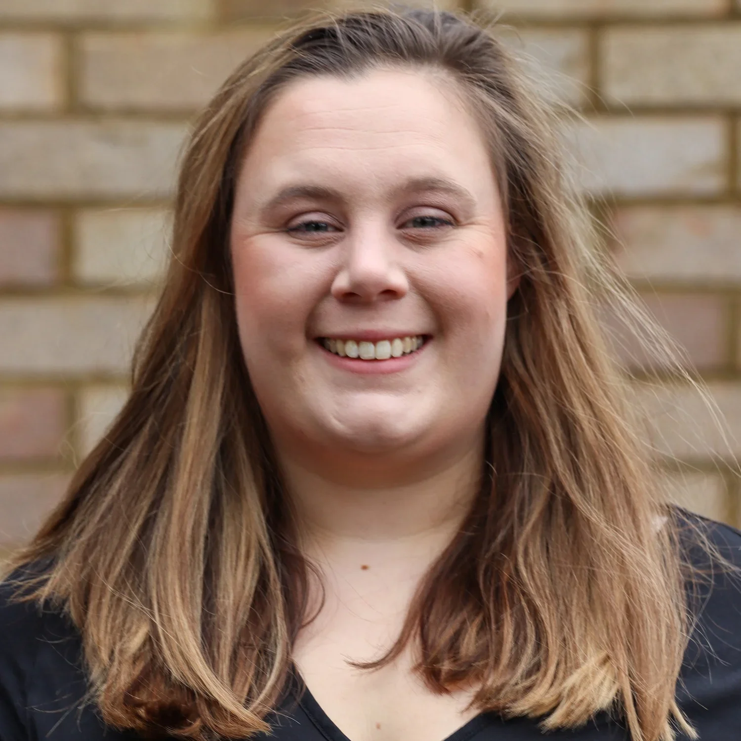 A woman with long, wavy, light brown hair smiling in front of a brick wall.