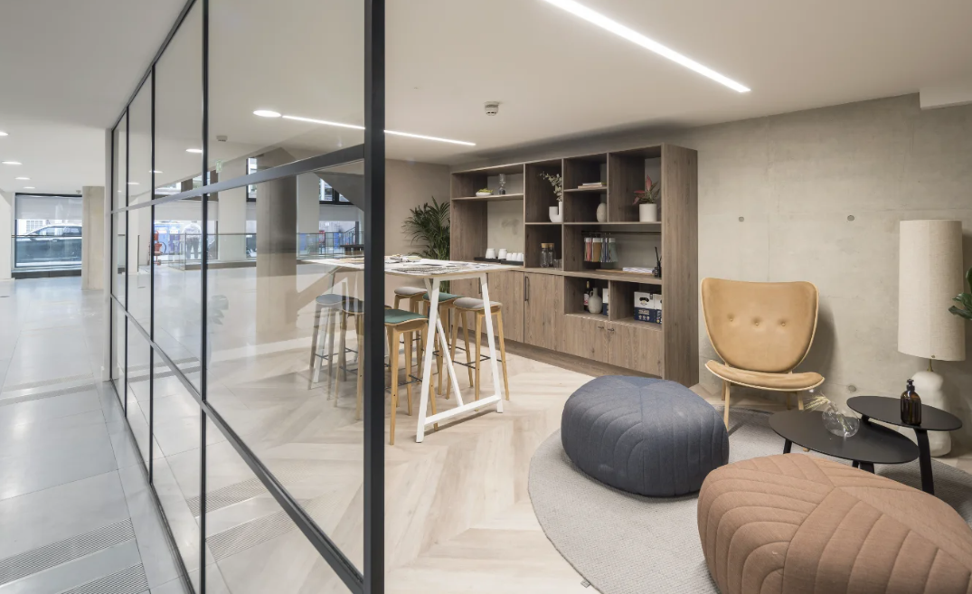 Modern office lounge with glass partitions, wooden bookshelf, beige and gray seating, and a small dining area with stools.