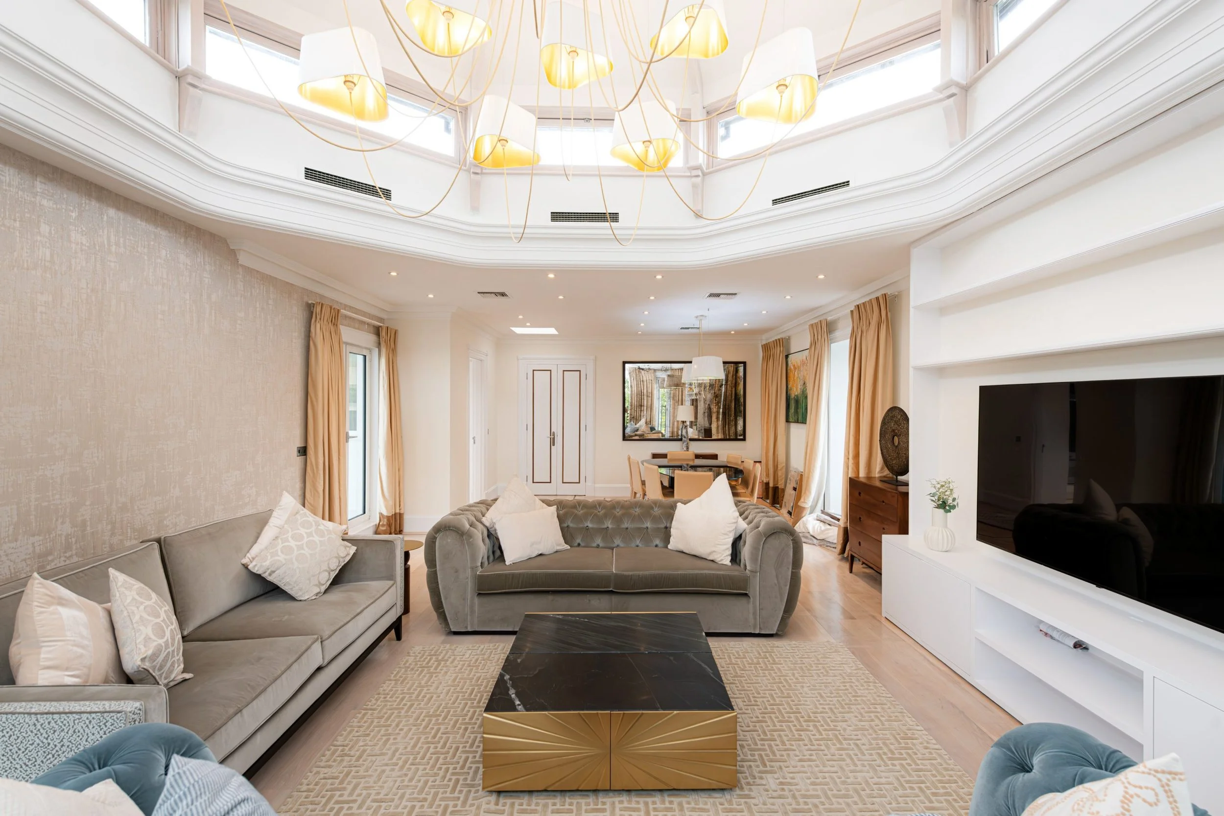 A spacious living room with beige walls, multiple windows with gold curtains, and a unique chandelier hanging from a white ceiling. The room contains a gray sofa, a tufted armchair, a black and gold marble coffee table, a large flat-screen TV, and a dining area with a round table and six chairs in the background.