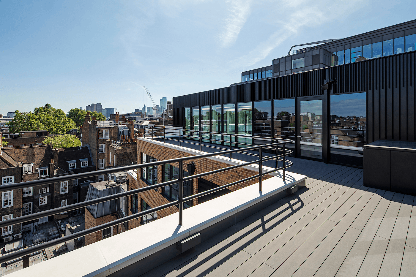 Rooftop terrace with glass and brick walls, metal railings, and an urban skyline with skyscrapers in the background under a blue sky.
