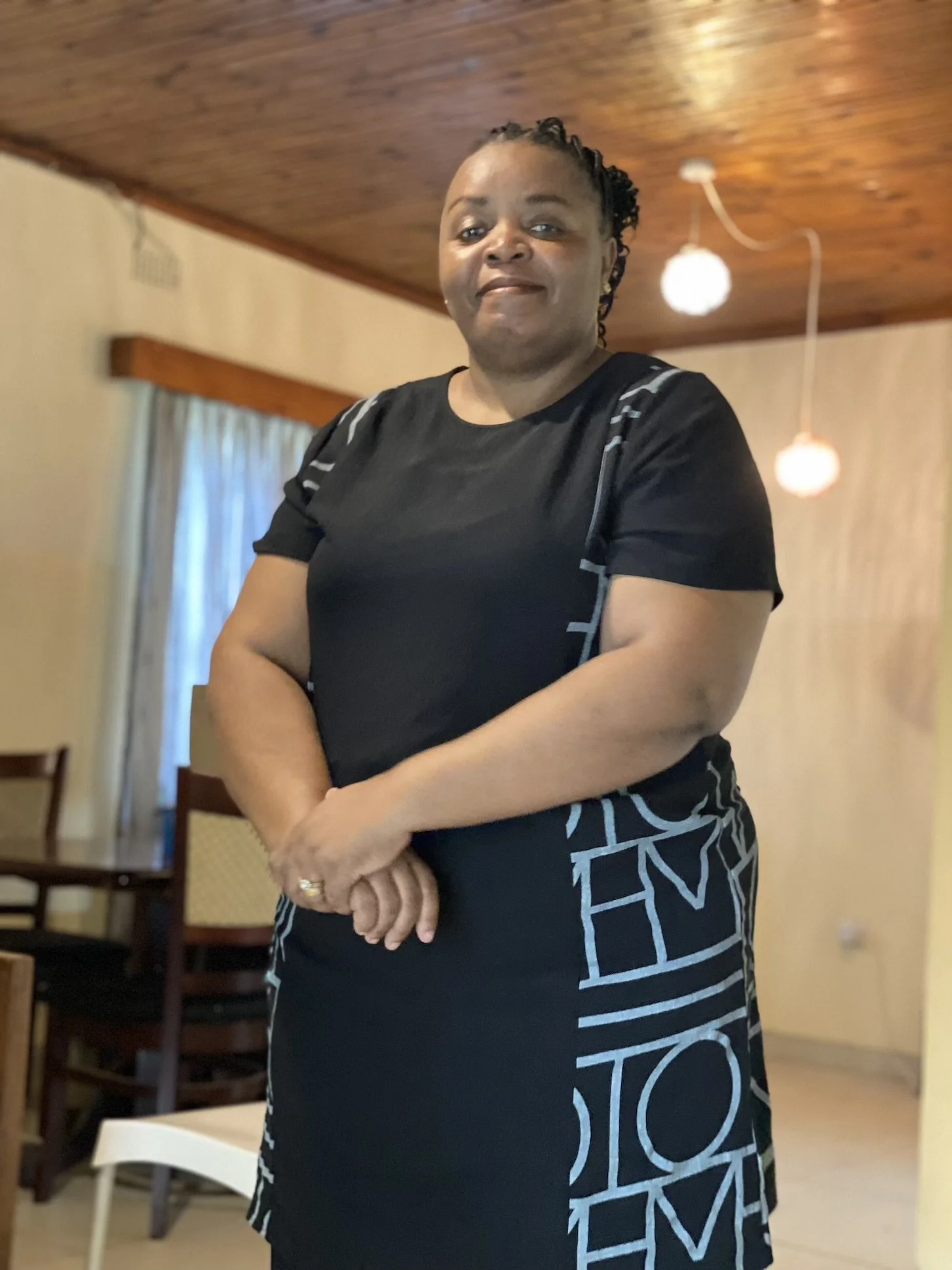 A woman standing indoors, wearing a black dress with geometric lettering, in a room with a wooden ceiling, hanging light fixtures, and a window with curtains.