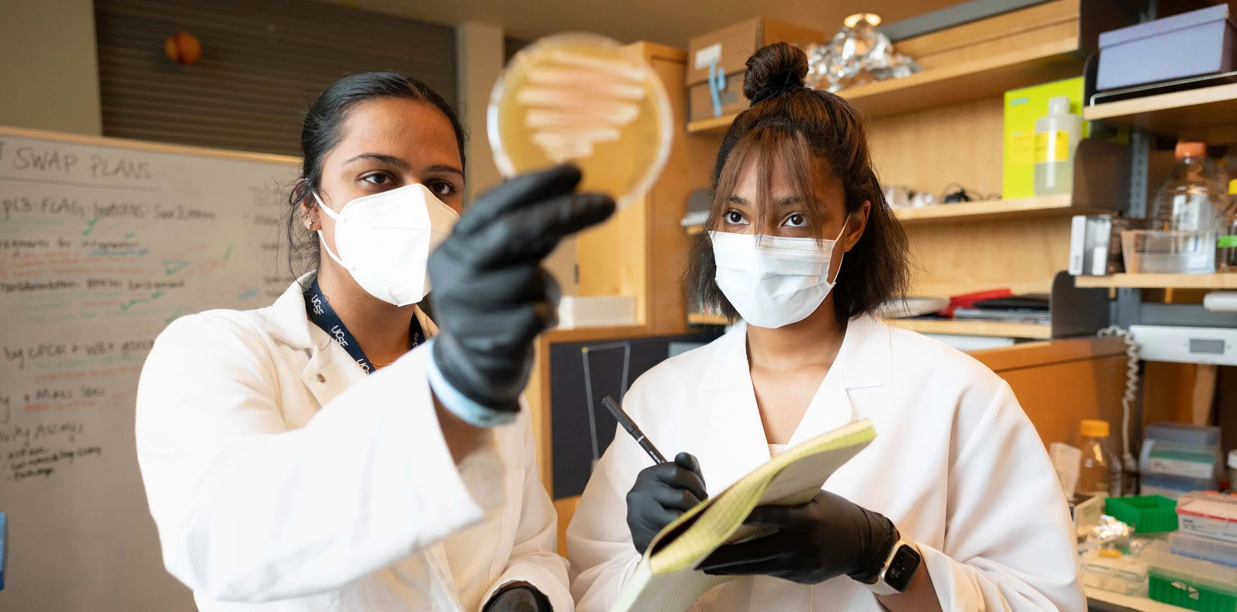 Two women scientists wearing white lab coats, face masks, and black gloves working in a laboratory. The woman on the left is holding a petri dish, and the woman on the right is writing in a notebook.