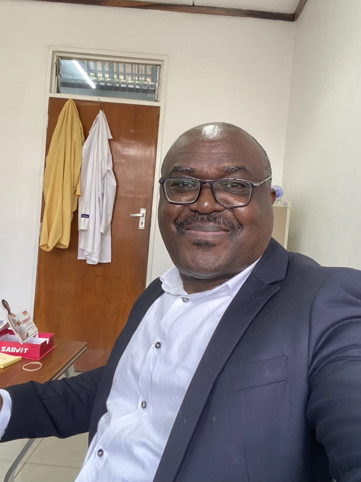 A man wearing glasses, a white shirt, and a dark blazer, smiling and taking a selfie in an indoor space, with a wooden door, a window, and hanging clothes in the background.
