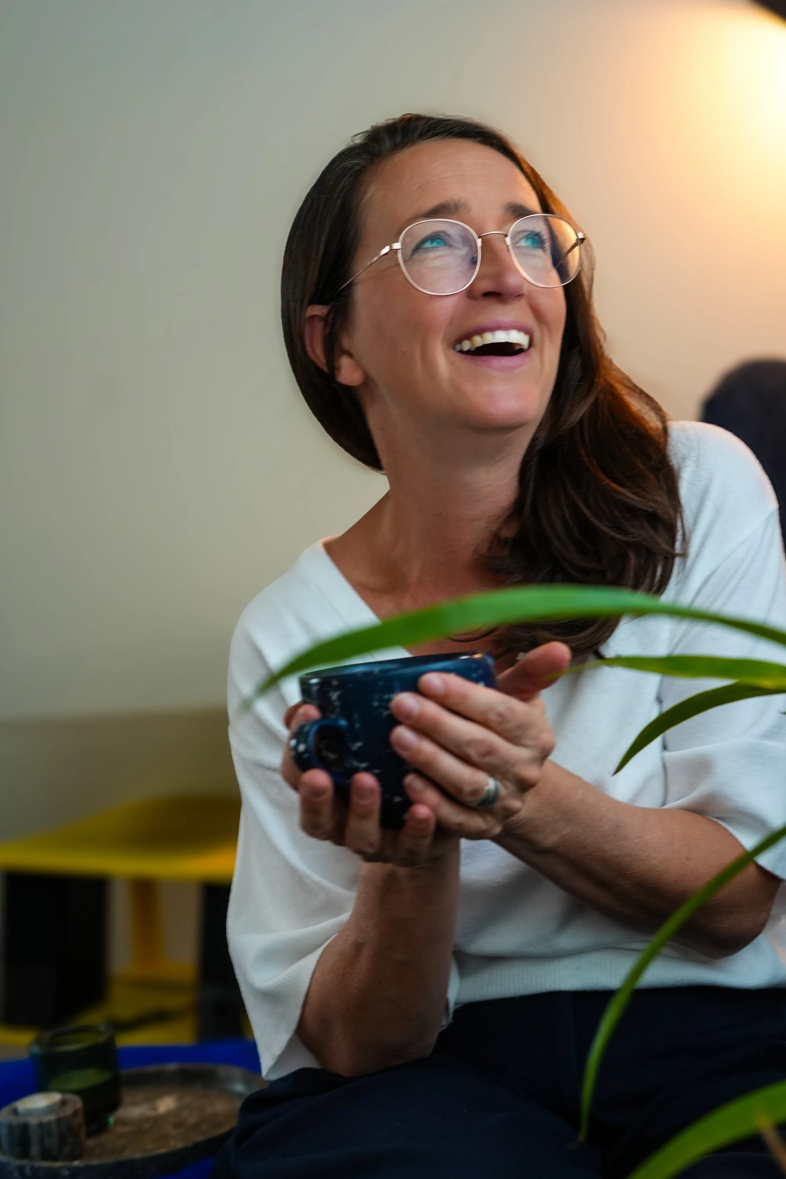 A woman with long brown hair, glasses, and a white top is smiling and holding a speckled blue mug.