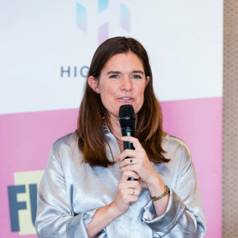 Woman with shoulder-length brown hair holding a microphone, speaking at an event with a pink and white background.