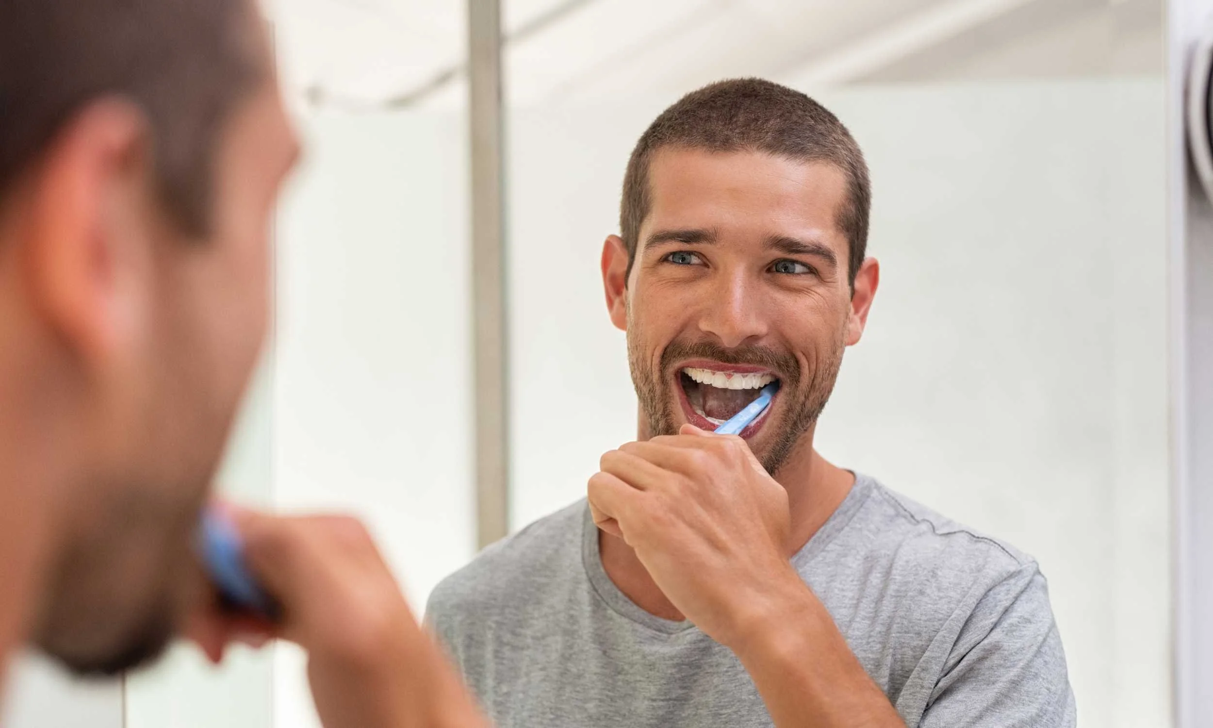 A man brushing his teeth while looking into a mirror, smiling.