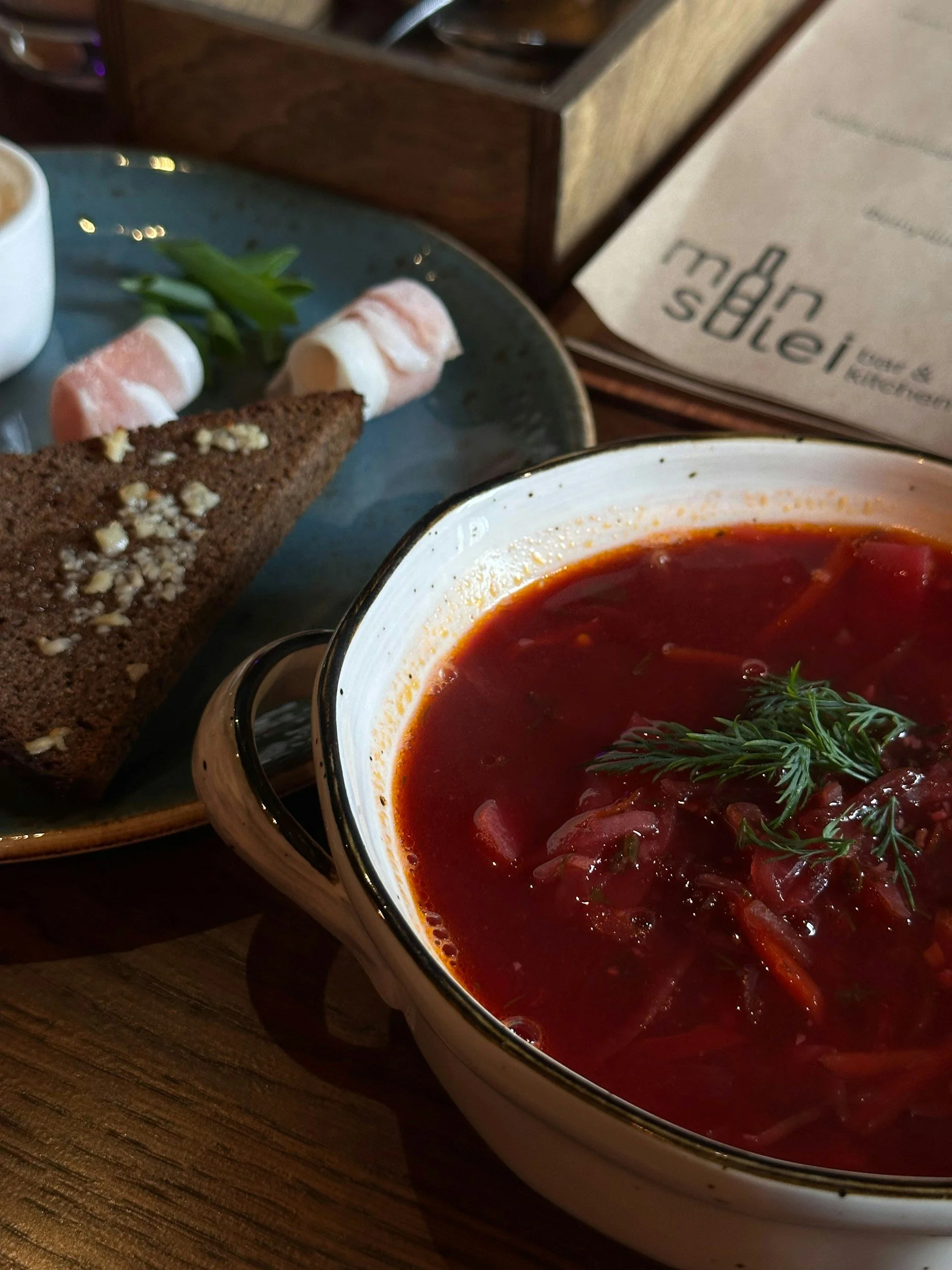 A bowl of borscht soup garnished with fresh herbs, with a slice of chocolate cake, a sliced roll, and a side dish on a dark wooden table.