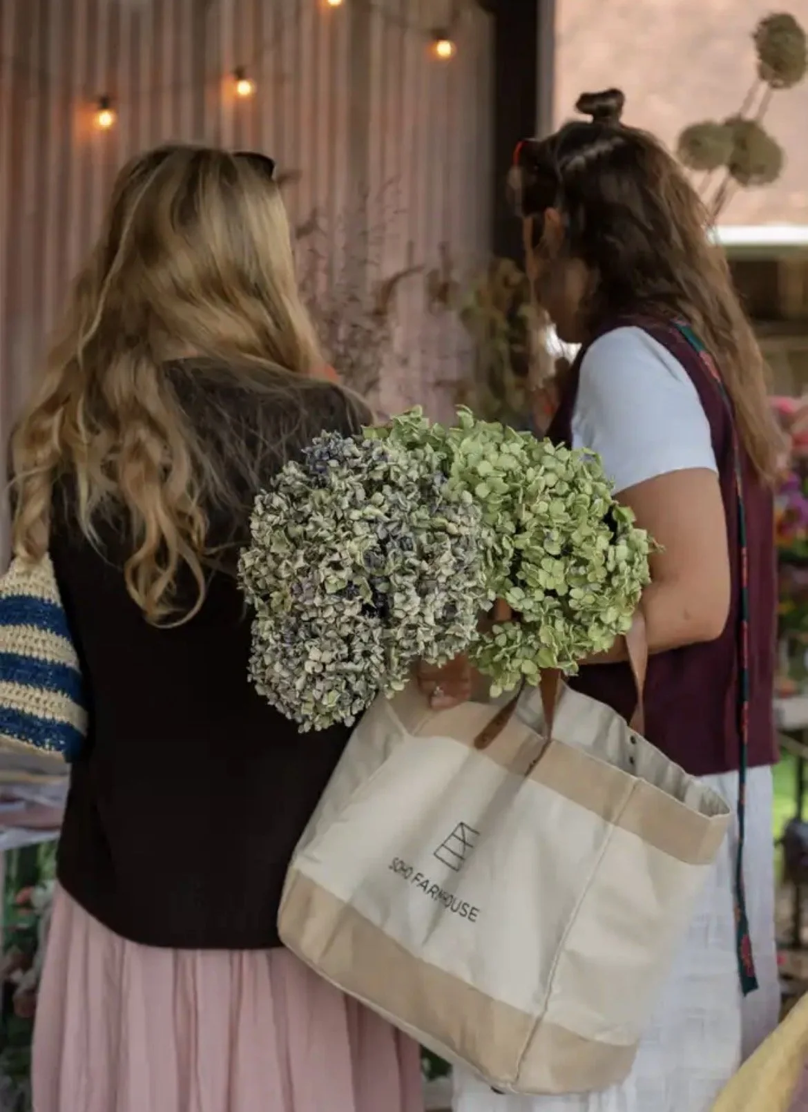 Two women shopping at a flower market, one holding a bouquet of white and purple flowers, the other holding a large beige tote bag with a flower shop logo, surrounded by flowers and plants with soft lighting in the background.