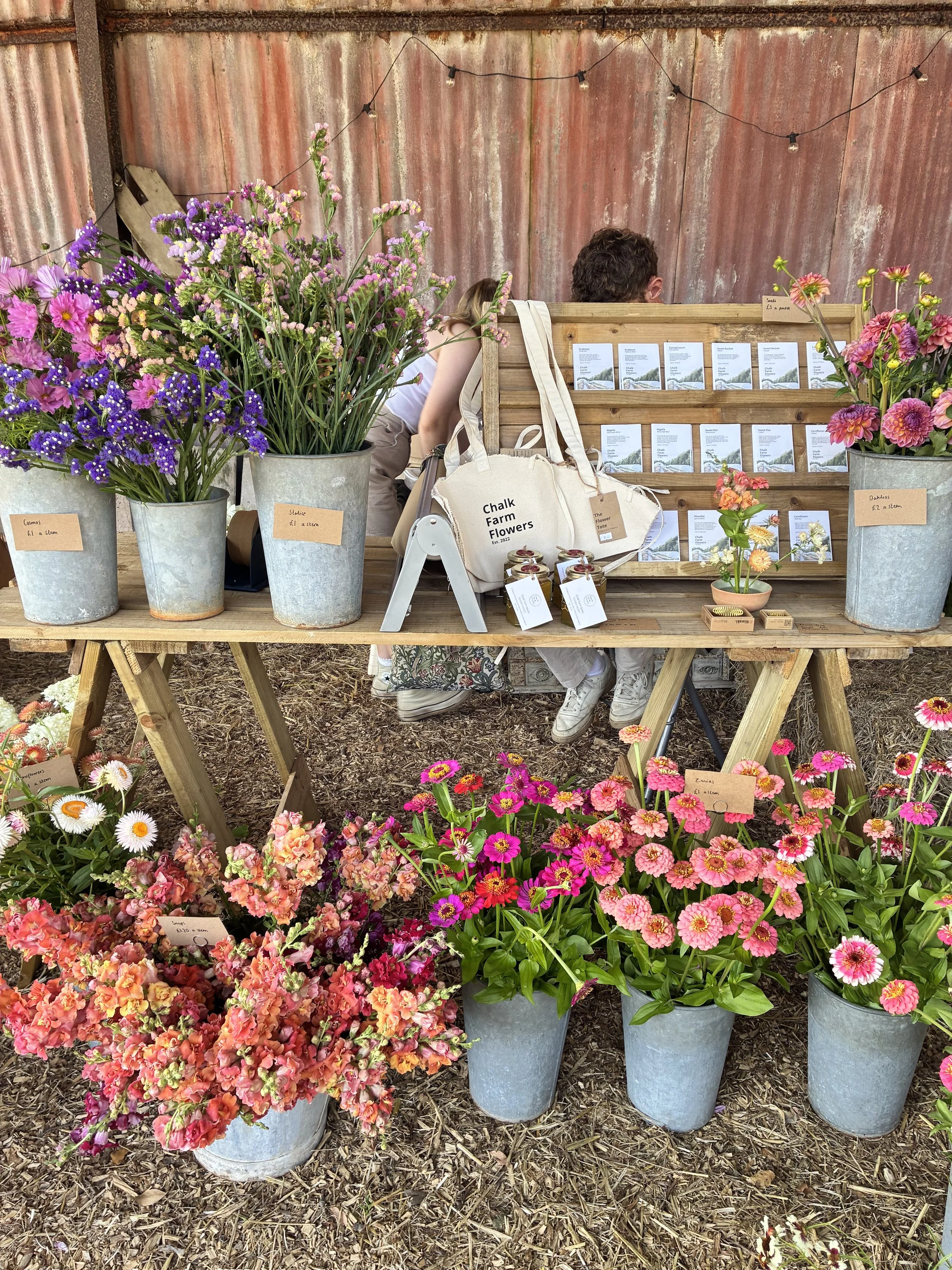 Display of colorful flowers in metal buckets at a flower stand at an outdoor market, with a rustic metal fence and string lights in the background. The flowers are labeled with small tags.