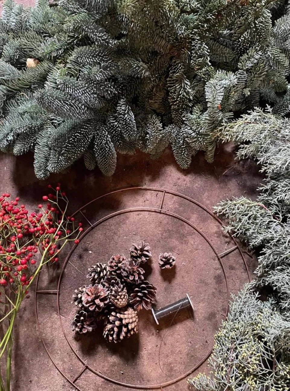 Arrangement of Christmas greenery, pinecones, and red berries on a surface with a circular metal frame and a small spool.