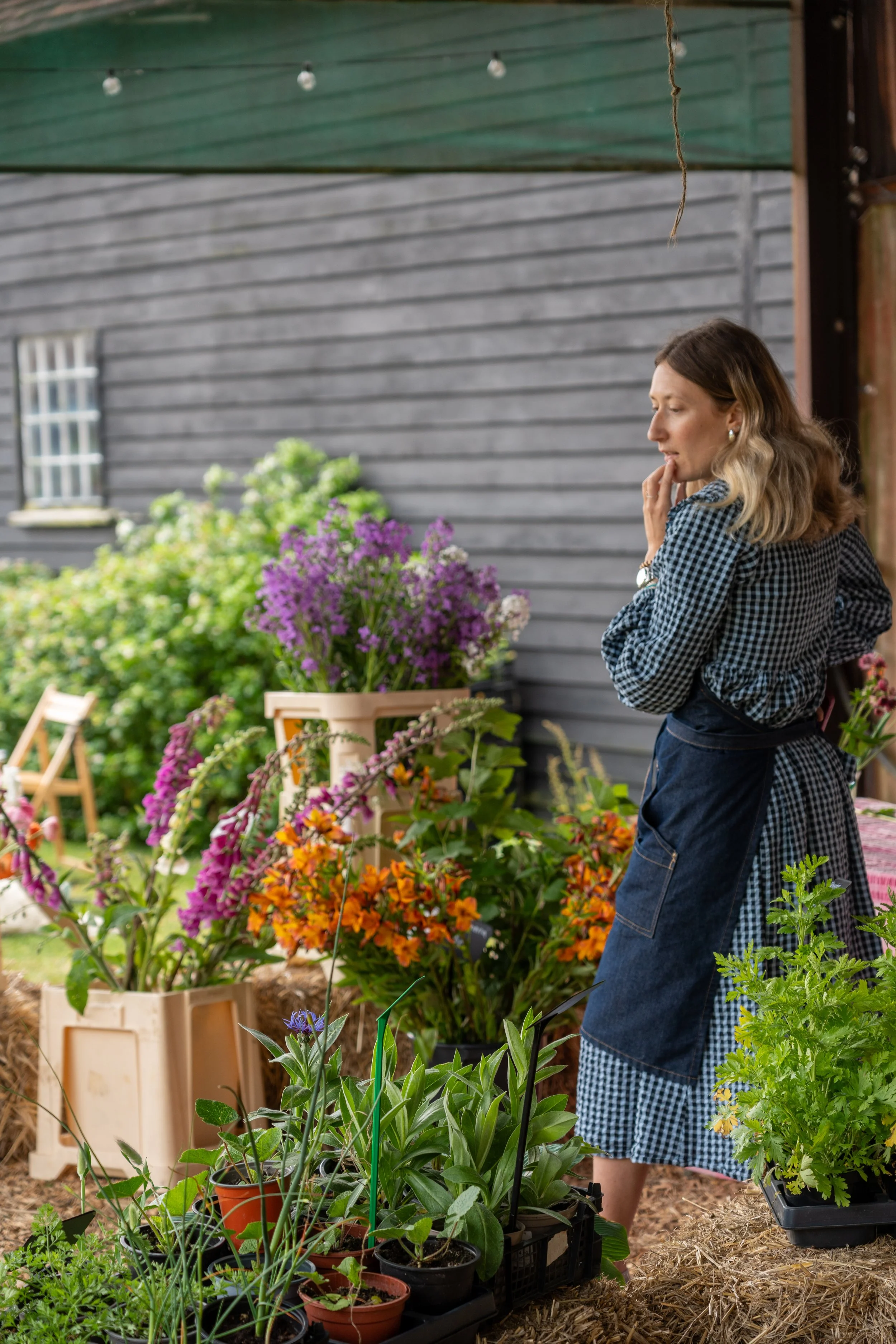Woman shopping at a plant nursery, browsing various potted plants and flowers with a gray barn wall and window in the background.