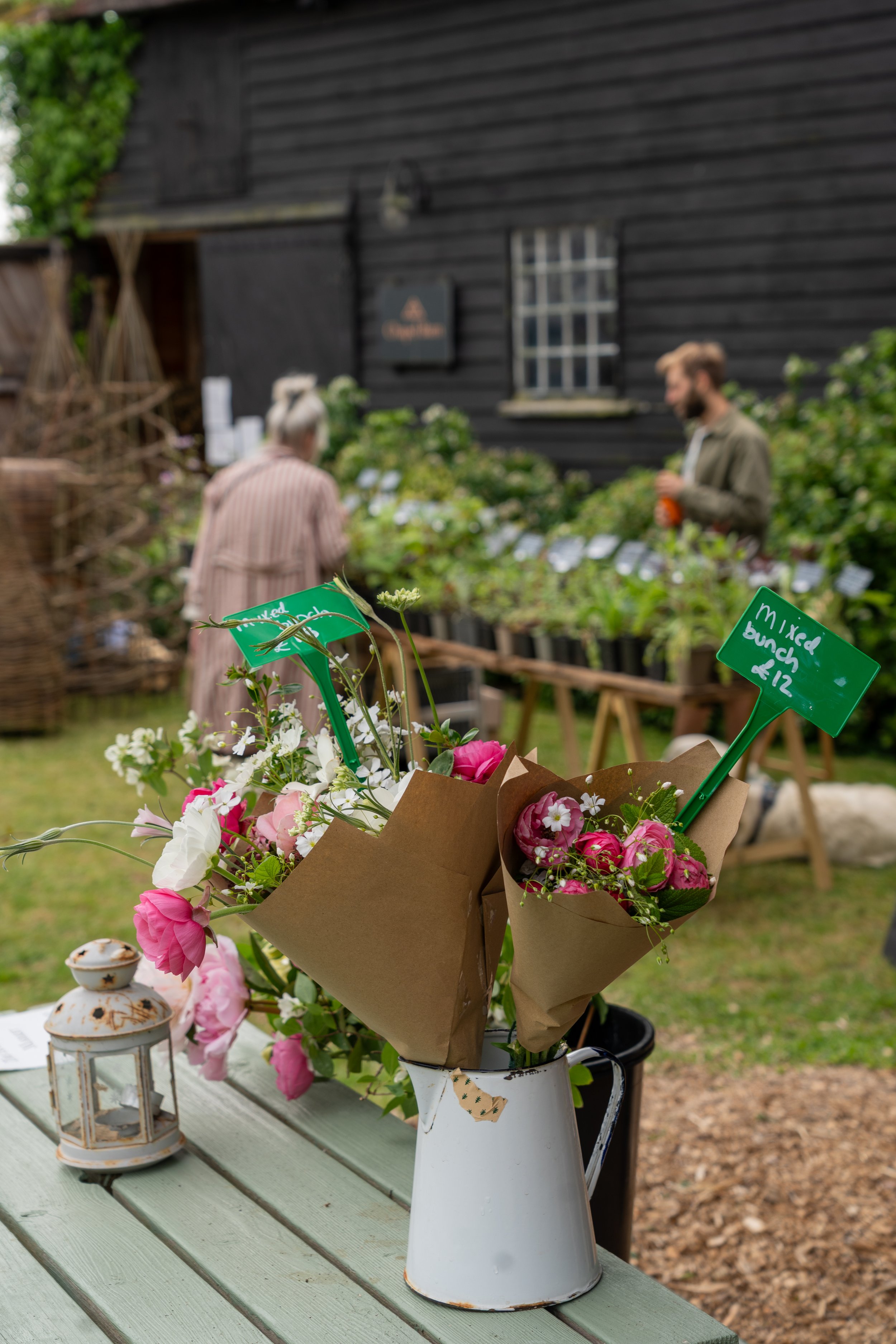 A bouquet of pink and white flowers in a white pitcher on a light green outdoor table, with a small lantern and decorative plants nearby, in front of a black wooden house with a window and a sign.
